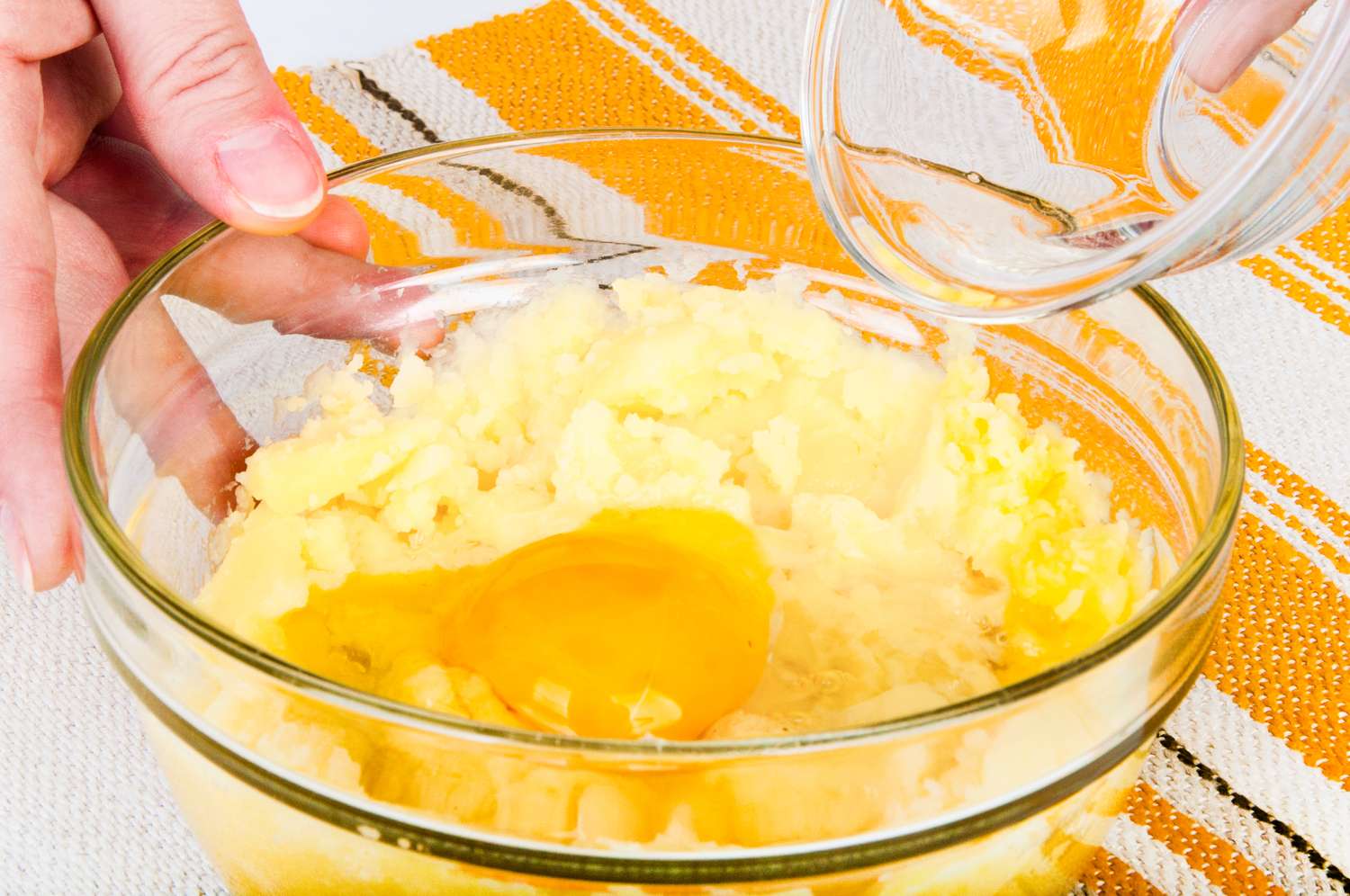 Mashed potatoes in a glass bowl with an egg yolk being mixed with a liquid being poured in from a smaller glass container