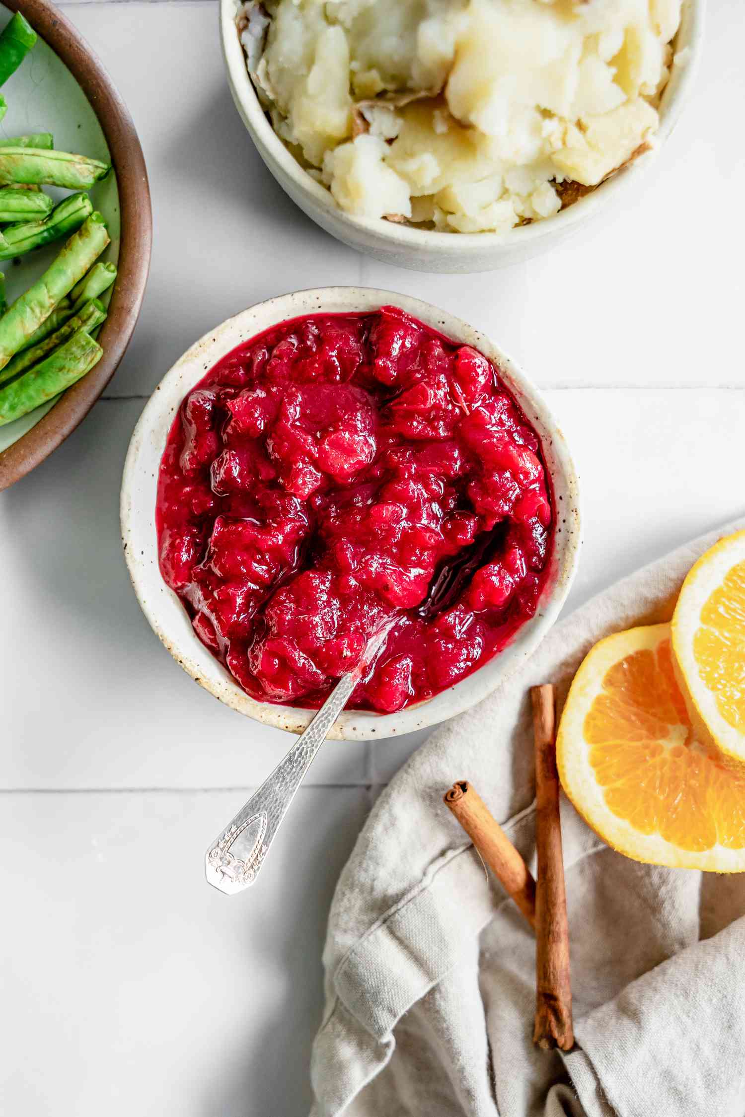 Instant Pot Cranberry Sauce Next to a Bowl of Mashed Potatoes, a Bowl of Green Beans, and Table Napkin with Orange Slices and Cinnamon Sticks