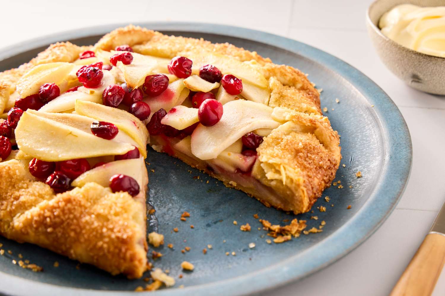 A pear and cranberry galette on a plate with a slice removed served next to a bowl of cream