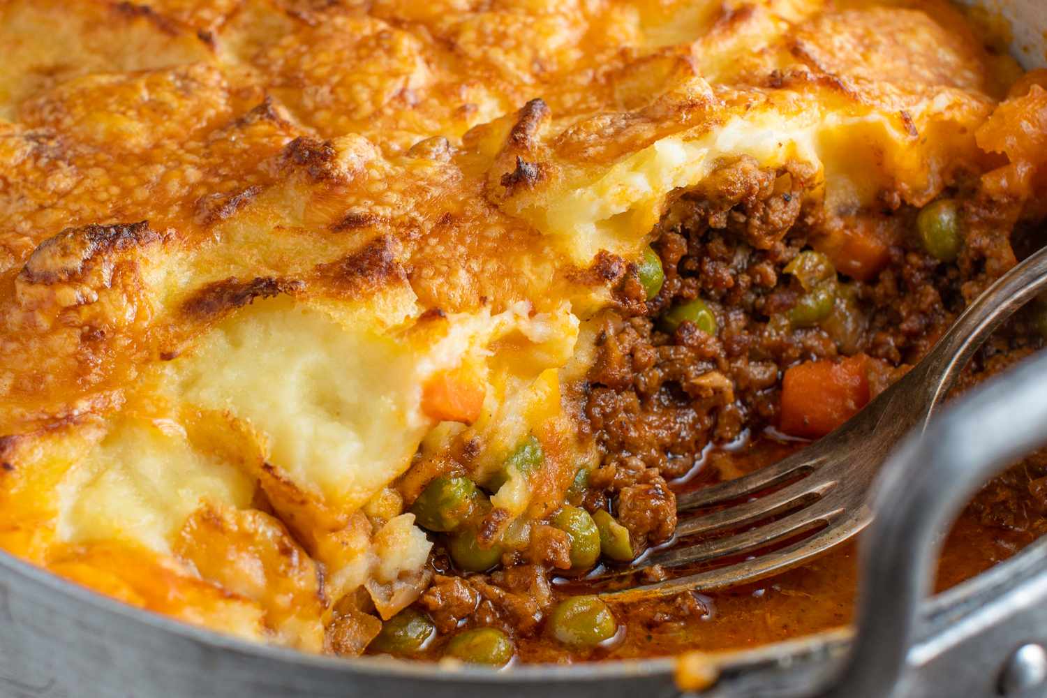 Shepherds pie with a fork in a serving in a pan showcasing layers of mashed potatoes beef peas and carrots