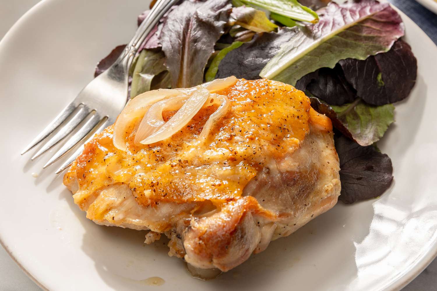 Overhead view of a white plate of a wine roasted chicken thigh topped with onions next to a bed of greens and a fork