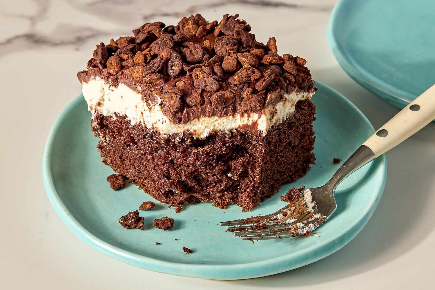 A slice of chocolate cake with cream topping and chocolate pieces on a plate with a fork