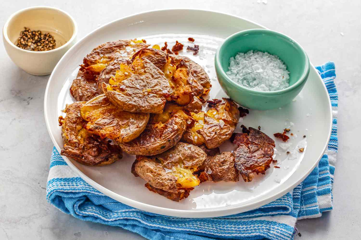 Smashed Potatoes and a Bowl of Flaky Slat on a Plate over a Kitchen Towel, and Next to It, a Small Bowl of Cracked Pepper