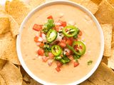 A bowl of queso dip garnished with tomatoes onions cilantro and jalapeo slices surrounded by tortilla chips