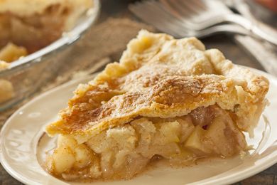 A slice of apple pie on a plate with a glass pie dish in the background