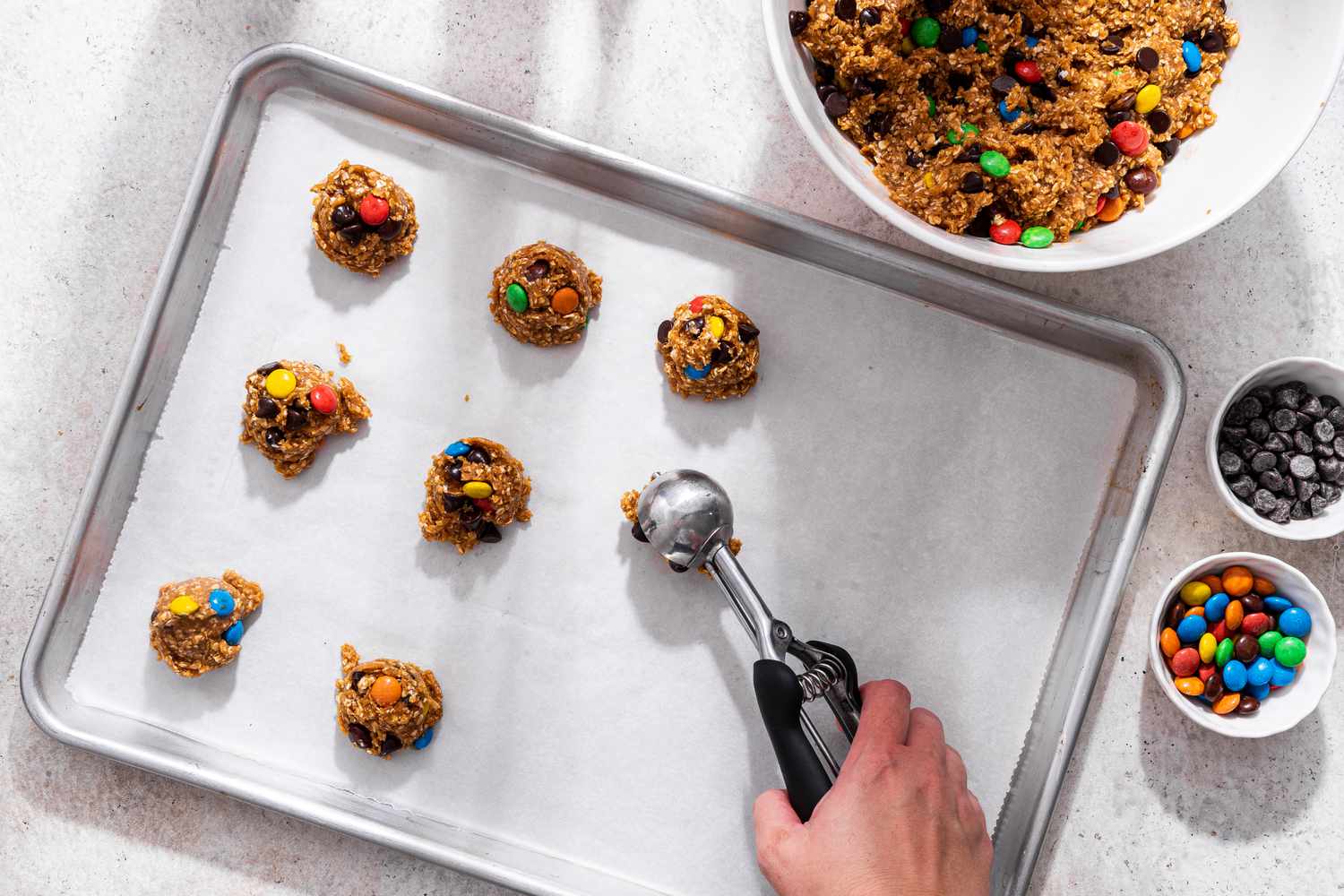 Dollops of monster cookie dough added to a parchment paper lined baking sheet using a cookie scoop, and in the surroundings, the bowl with more monster cookie dough and two small bowls with more mix-ins (one with chocolate chips, the other with M&M's)