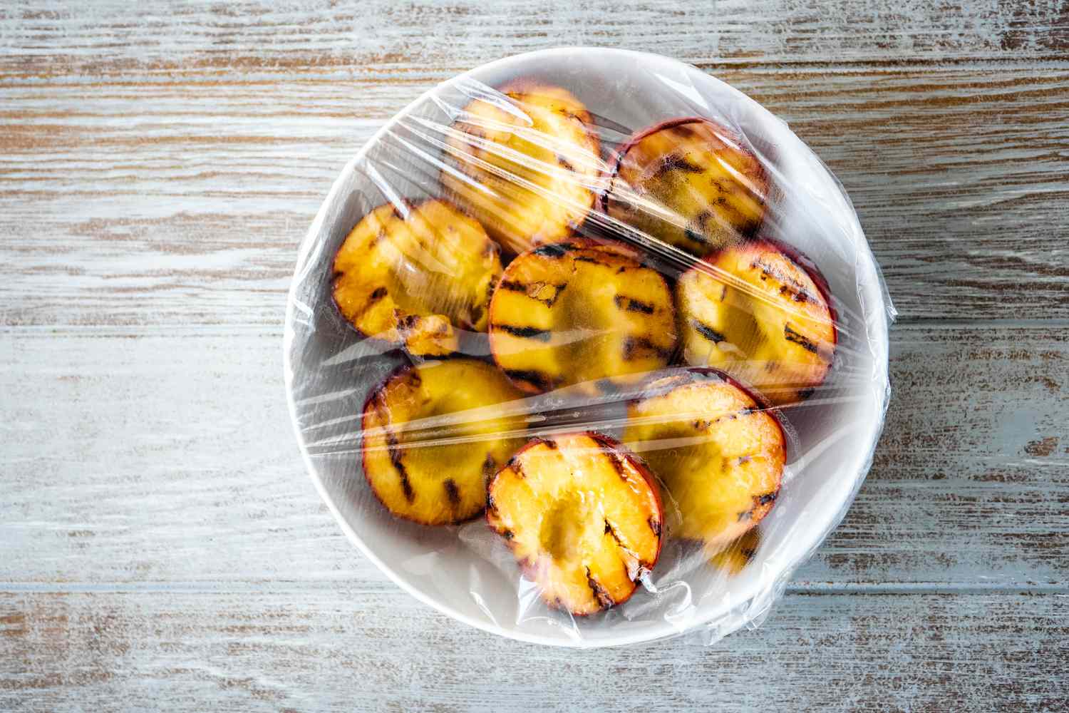A large bowl with grilled peaches covered in plastic wrap to make grilled peach pitcher margaritas.