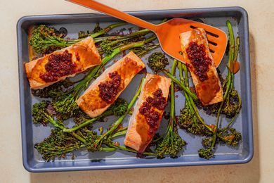 Overhead view of a baking sheet of four cooked salmon filets topped with chili crisp and stems of broccolini with an orange spatula under a piece of salmon