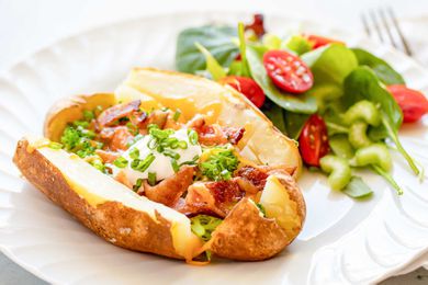 Cheesy, broccoli, bacon loaded baked potato on a white plate with a salad of spinach and tomato behind it on a white plate. The potato is sliced down each side and the top and bottom. Chopped bacon, broccoli, scallions and shredded cheddar cheese are on the potato. A spoonful of sour cream is in the middle of the potato. A fork is to the right of the plate.