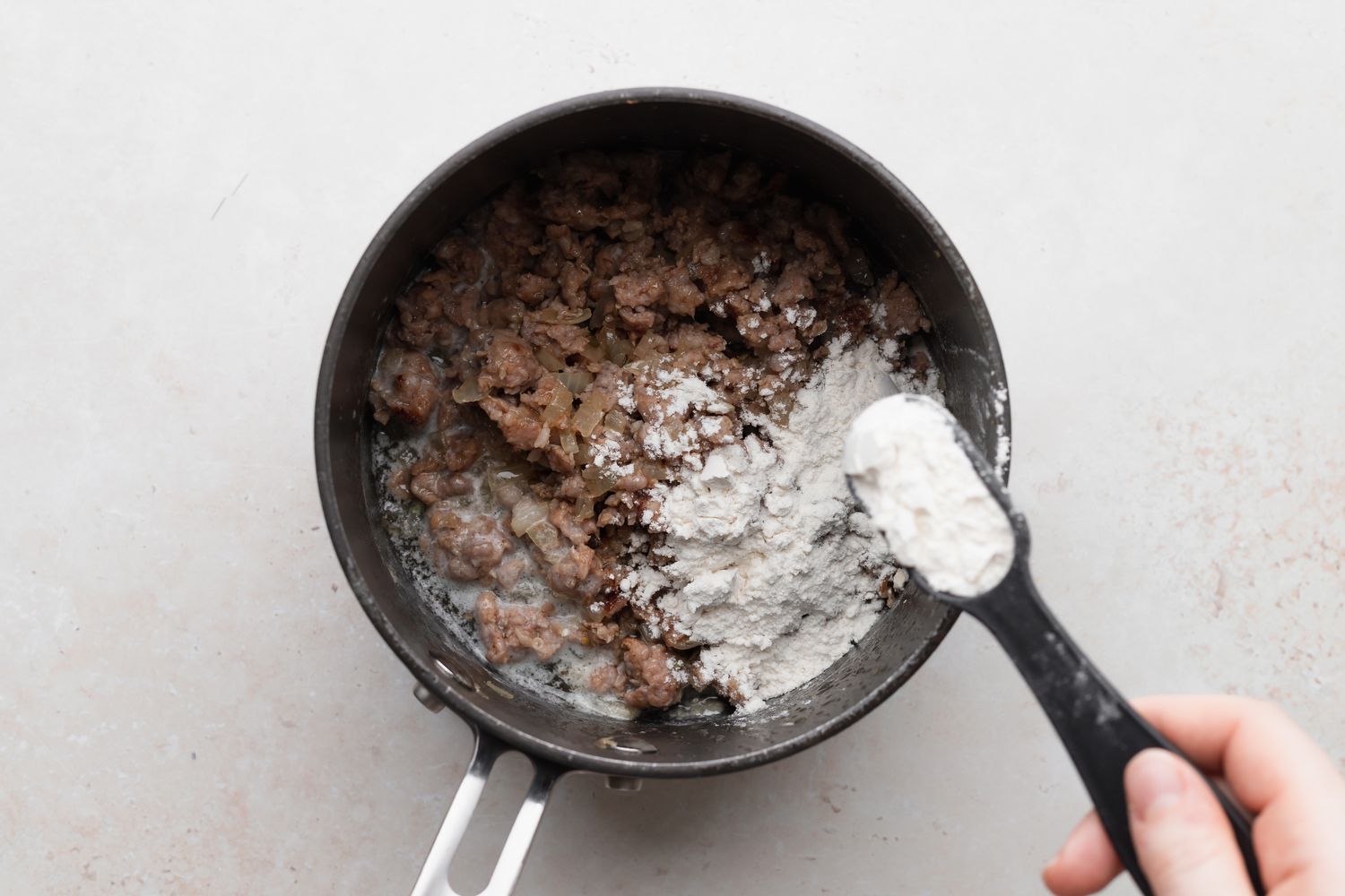 Adding flour to a pot with sausage to show how to make biscuits and gravy.