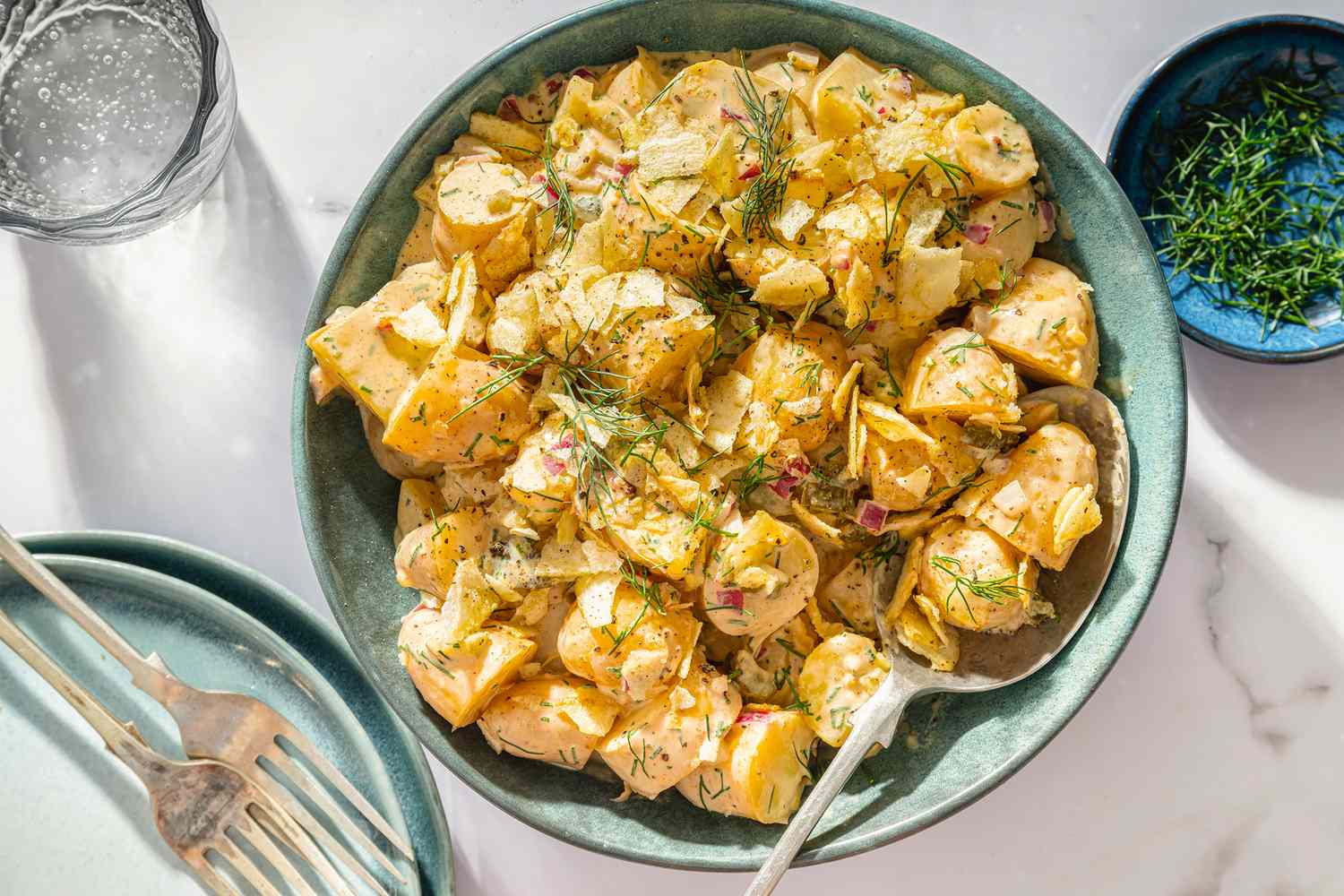 A bowl of dill pickle potato salad with a spoon garnished with fresh dill placed on a table alongside plates and flatware