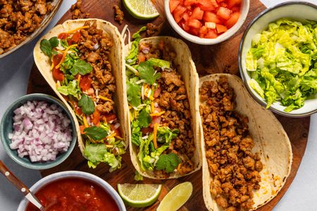Three tacos with sausage filling on a plate, surrounded by bowls of taco toppings including diced tomatoes, lettuce, onions, and salsa