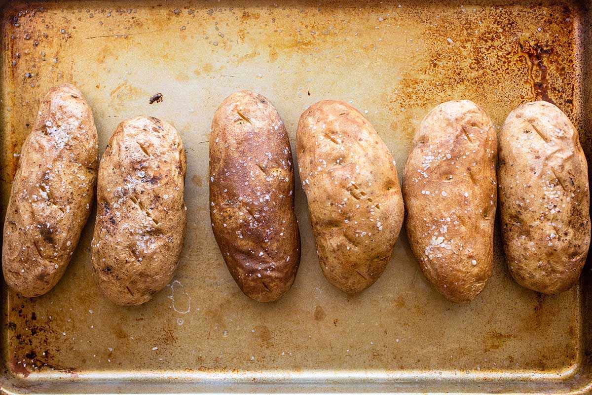 Six russet baked potatoes are on a worn baking sheet. The skin is dimpled and salt covers each one.