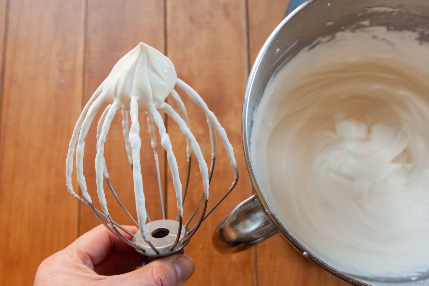 Whipped cream frosting with cream cheese in a mixer bowl and a person holding the whisk attachment next to the bowl.