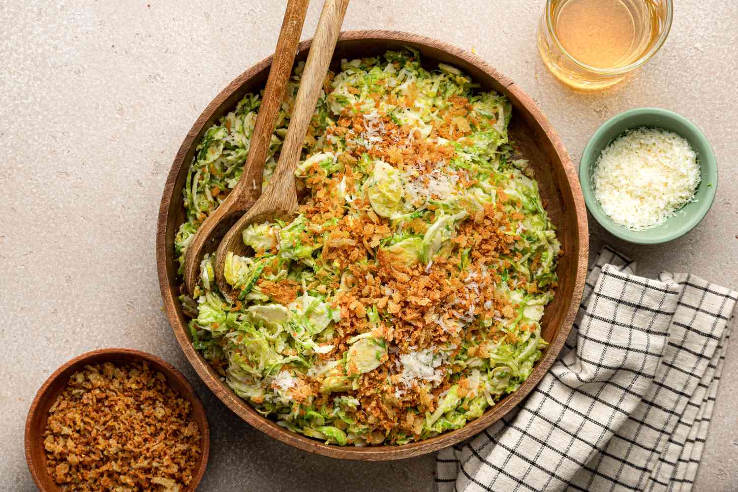 Bowl of Brussels Sprouts Caesar Salad with Lemony Breadcrumbs Next to Serving Utensils on a Table Napkin, a Bowl with More Toppings, a Bowl with Shredded Parmesan, and a Glass