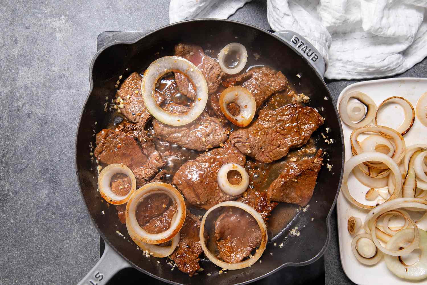 Bistek Cooking in a Cast Iron Skillet With Some of the Onion Slices After Being Flipped, and Next to the Cast Iron Skillet, a Plate With Cooked Onions and a White Kitchen Towel