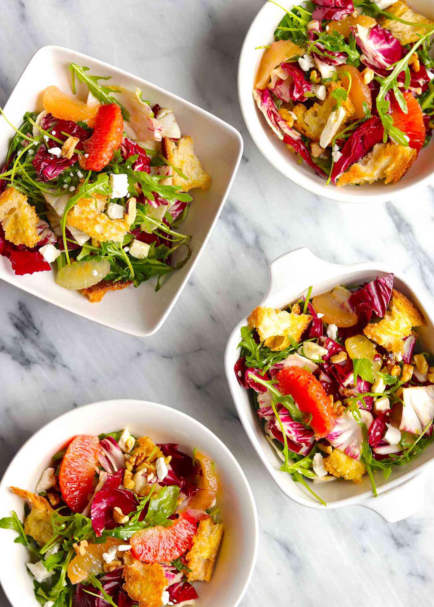 Overhead view of four white bowls with winter salad inside. Arugula, radicchio, supremed citrus, croutons and crumbled feta are visible in each bowl.