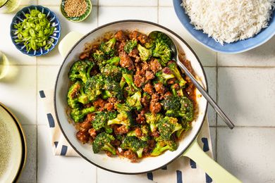 Bowl of ground beef and broccoli stir fry at a table setting with a bowl of rice, bowl of sesame seeds and a glass of water