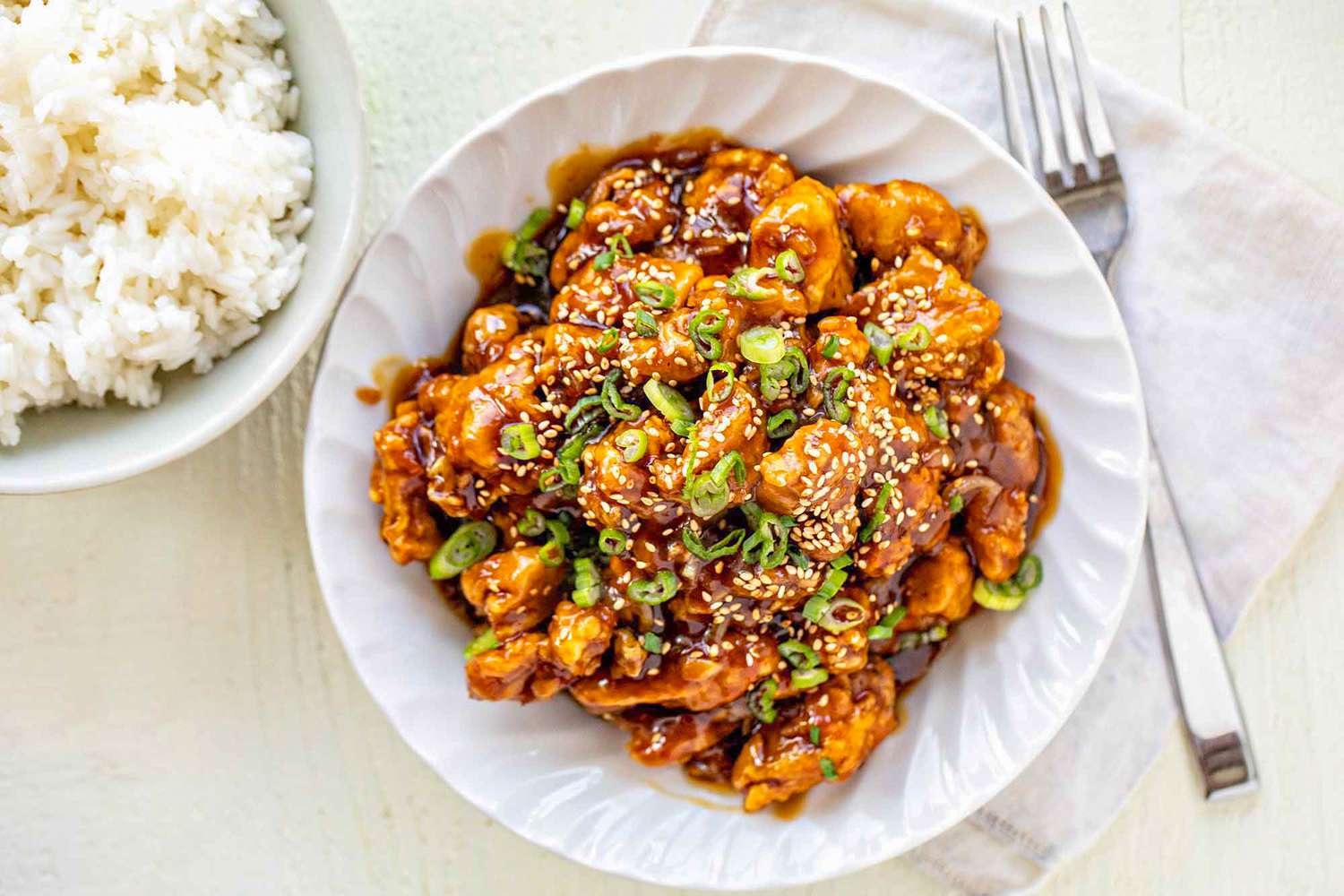 Orange Chicken in a Bowl Next to a Bowl of Rice, a Table Napkin, and a Fork