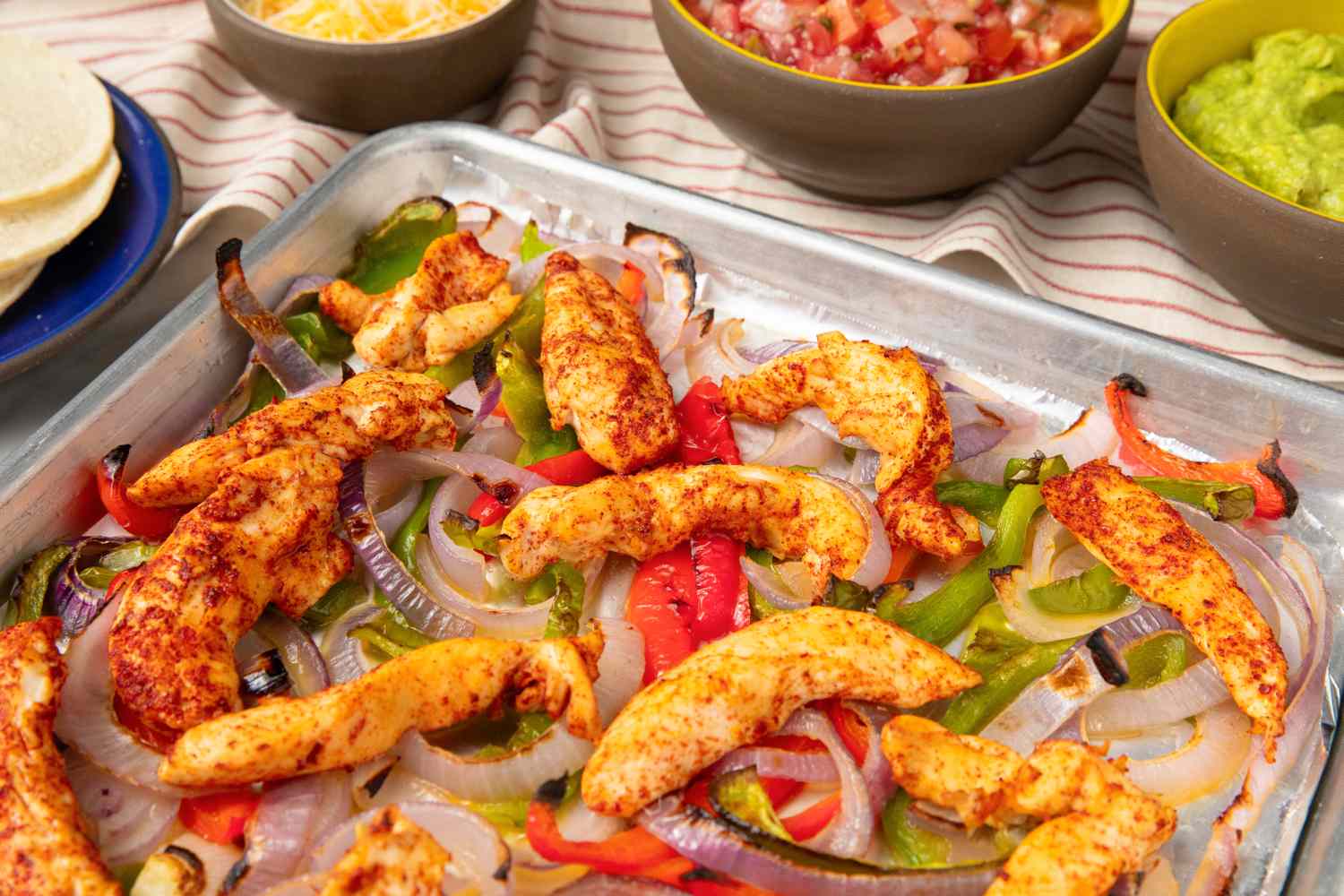 Sheet pan with cooked chicken strips and assorted vegetables surrounded by bowls of tortilla shells shredded cheese salsa and guacamole