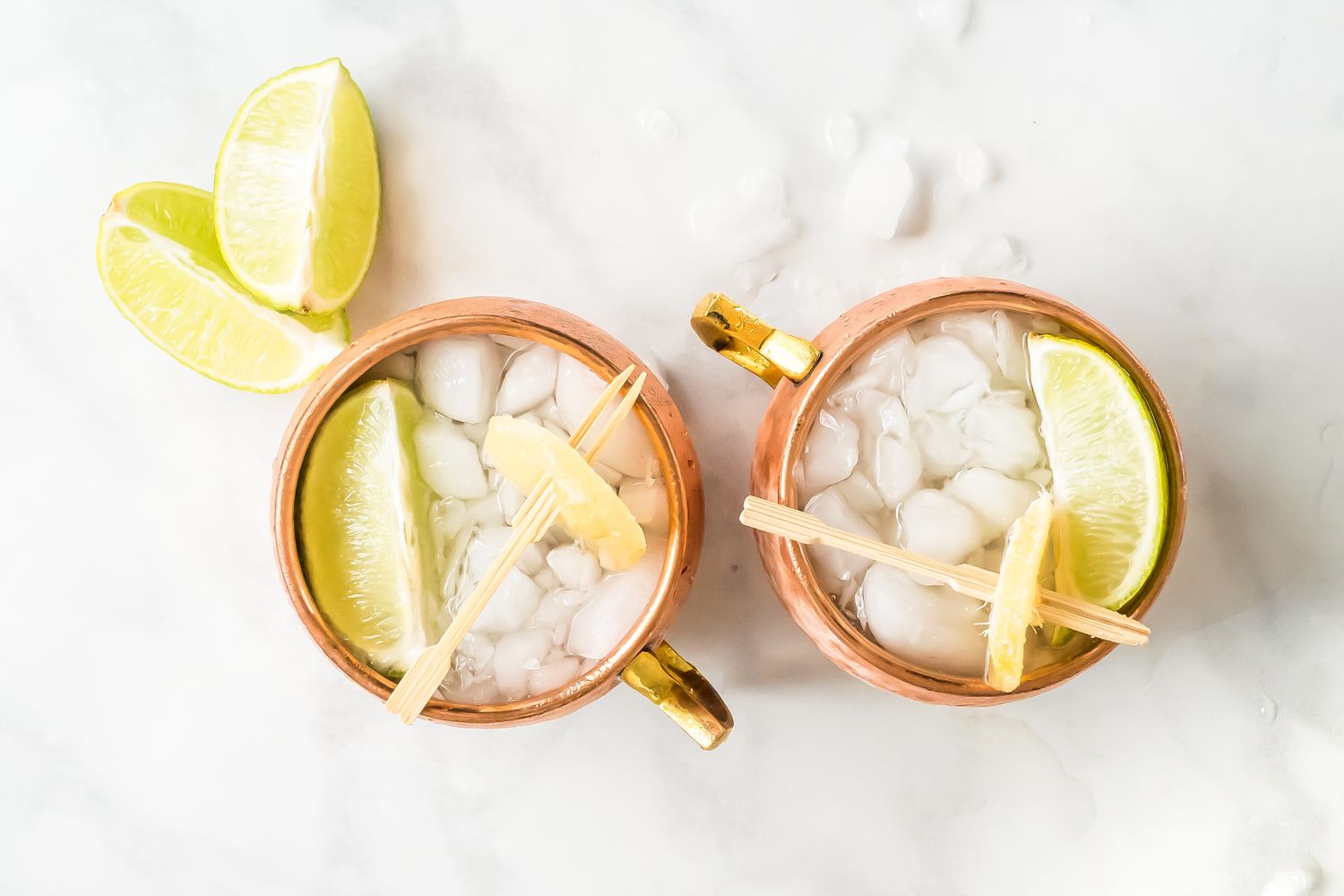 Two Copper Mugs of Mezcal Mule Garnished with Lime Wedges and Candied Ginger, Next to More Lime Wedges