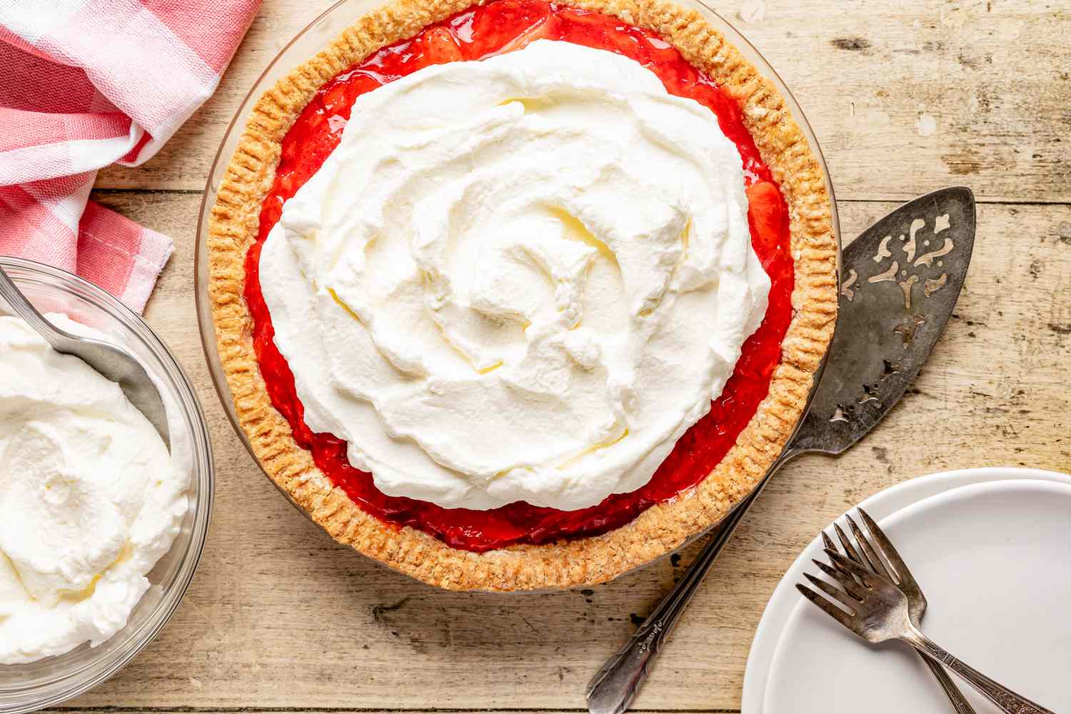 fresh strawberry pie at a rustic table with a bowl of whipped cream, a stack of plates, and a kitchen towel