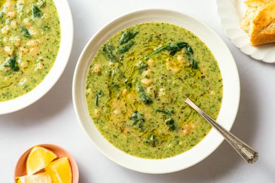 Overhead view of a white bowl of white bean and spinach soup with a spoon next to a plate of bread and bowl of lemon slices