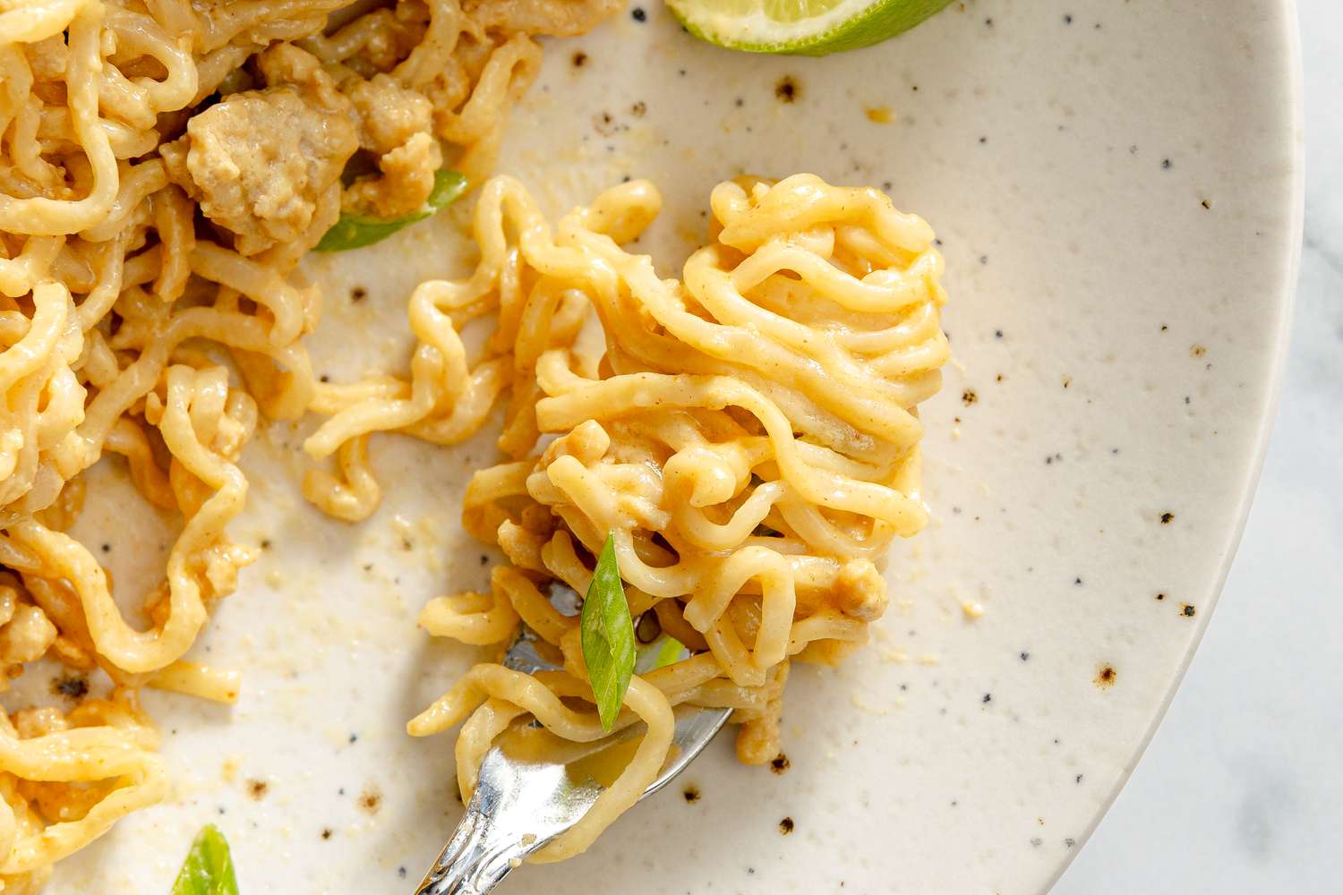 A plate of peanut chicken noodles with garnish and a piece of lime on the side, fork twirling a portion of noodles