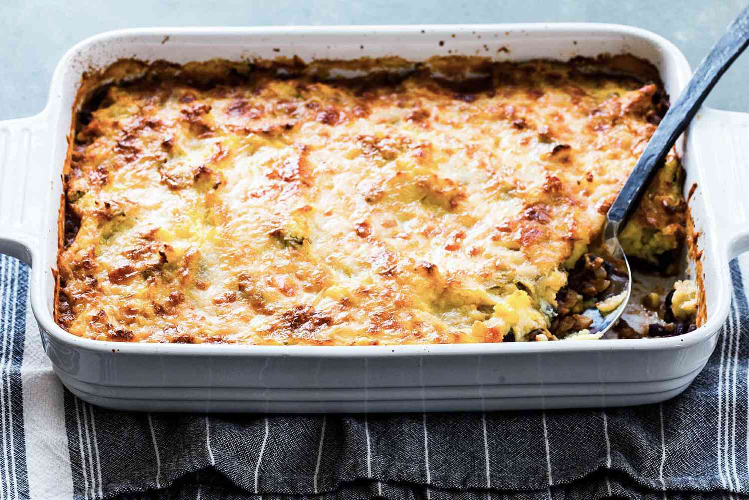 Horizontal view of easy shepherd's pie with a striped dish towel underneath white casserole dish. Serving spoon insdie with a scoop missing.