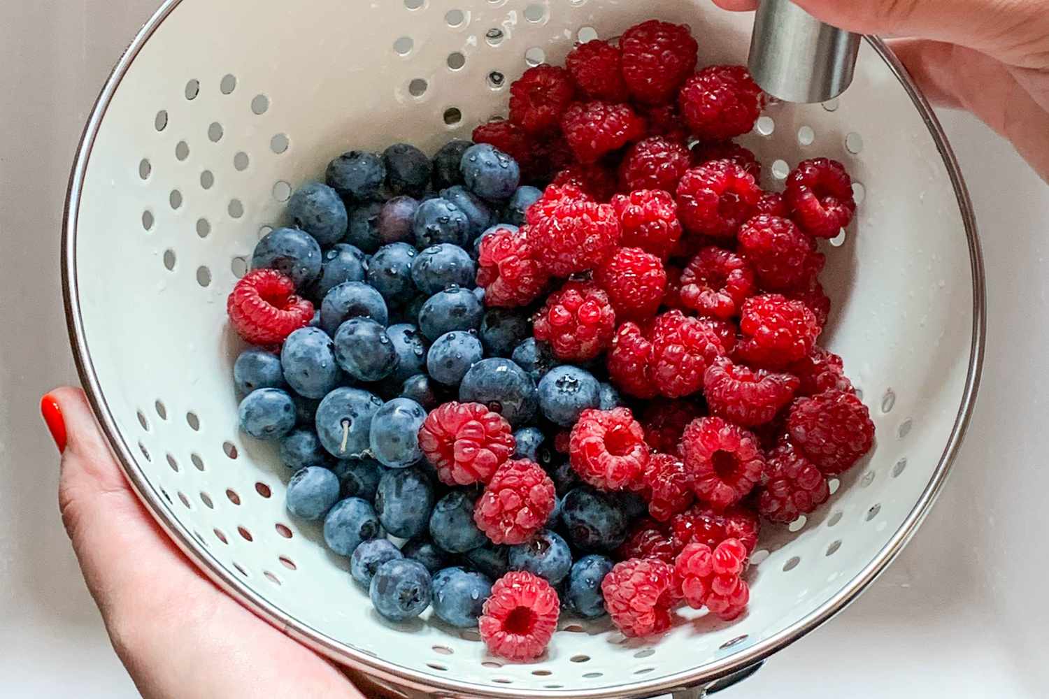 Hands holding a colander as they rinse strawberries and blueberries