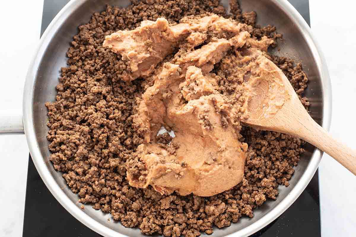 Refried beans being stirred into cooked ground beef with a wooden spoon in a pan