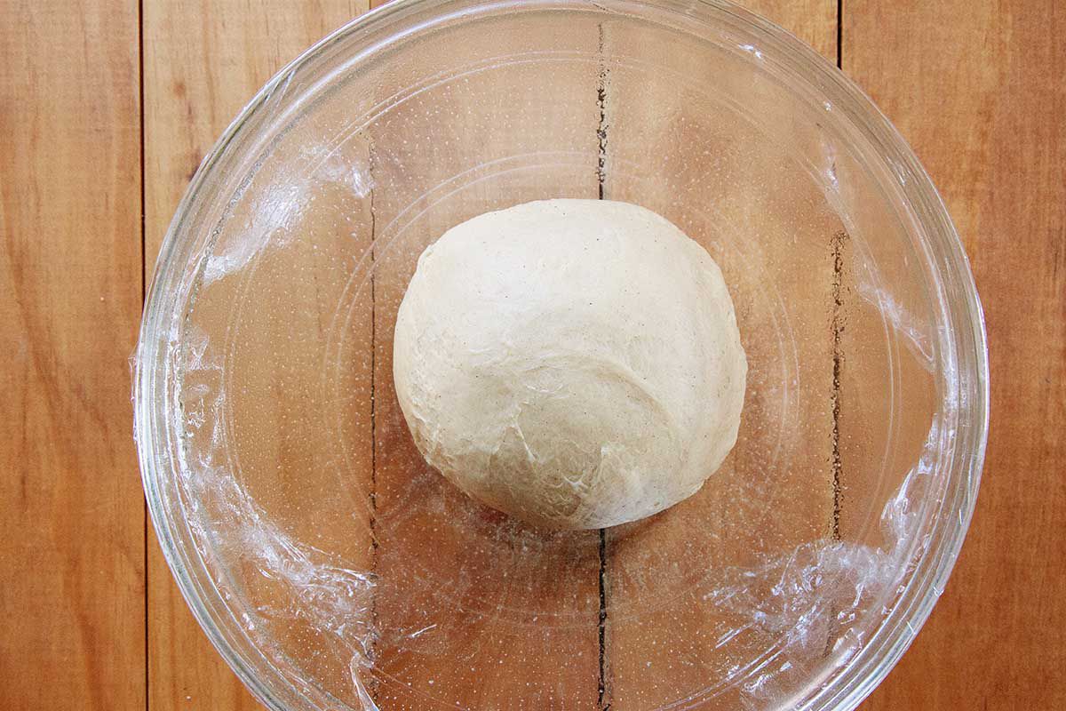 Dough for homemade king cake is resting in a large glass bowl and covered with plastic wrap.