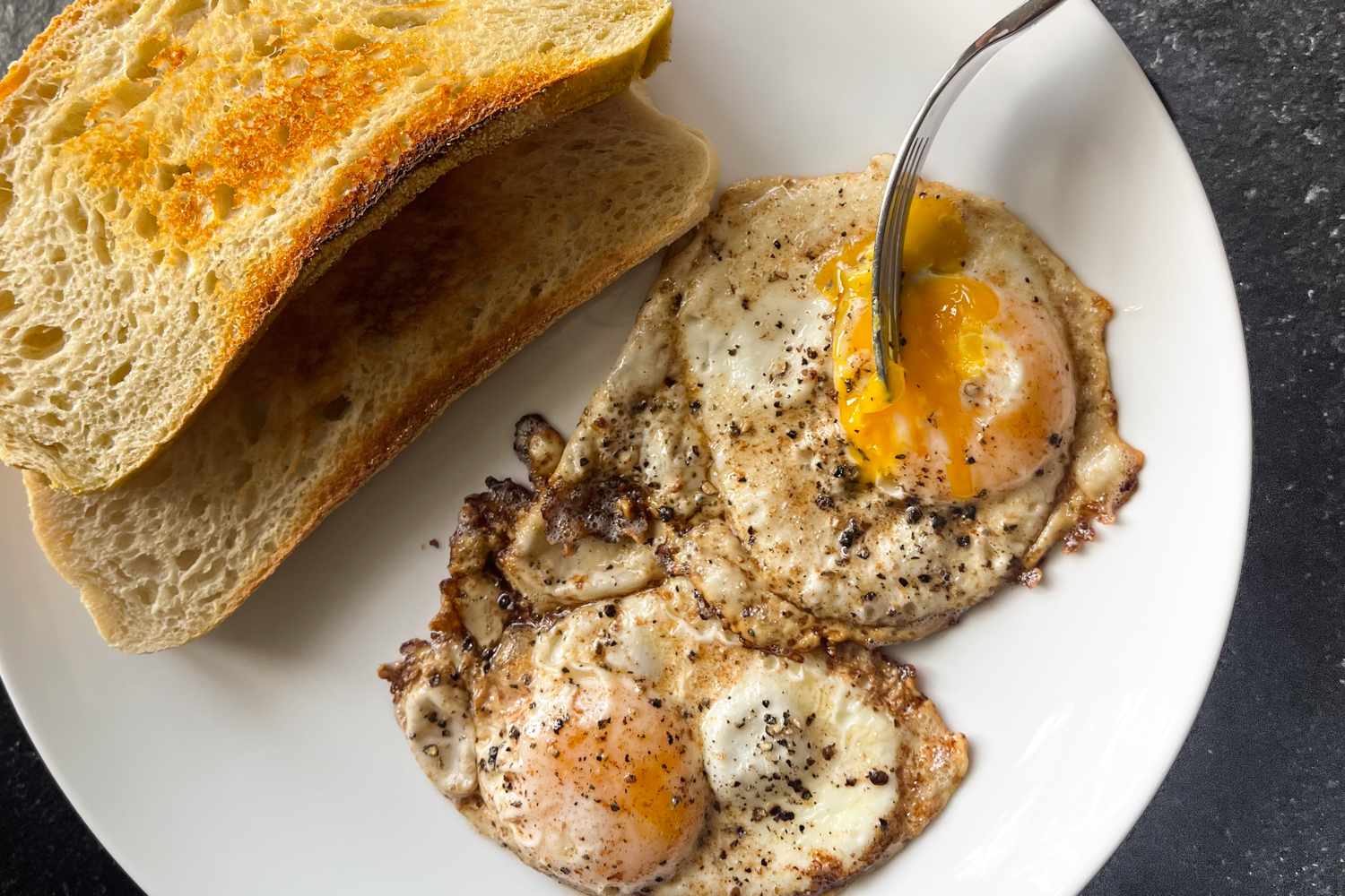Fried eggs with cracked pepper on a white plate served with slices of toasted bread and a fork breaking into one egg yolk
