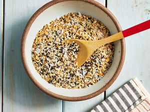 Bowl of Everything Bagel Seasoning With a Wooden Spoon and Next to a Black and White Stripped Kitchen Towel