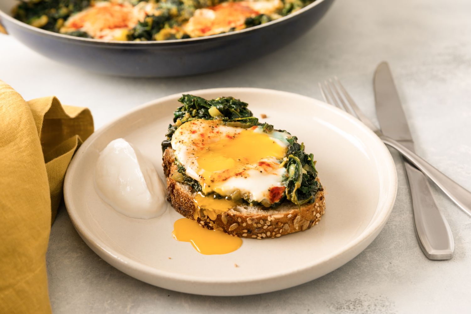 A plate with bread topped with sauted spinach an egg and a side of yogurt with a skillet in the background