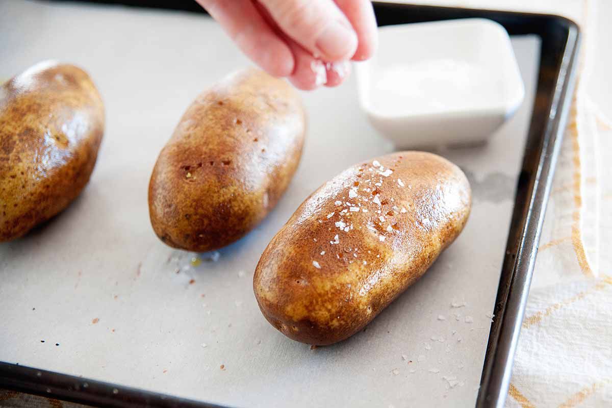Three russet potatoes being salted on a baking sheet