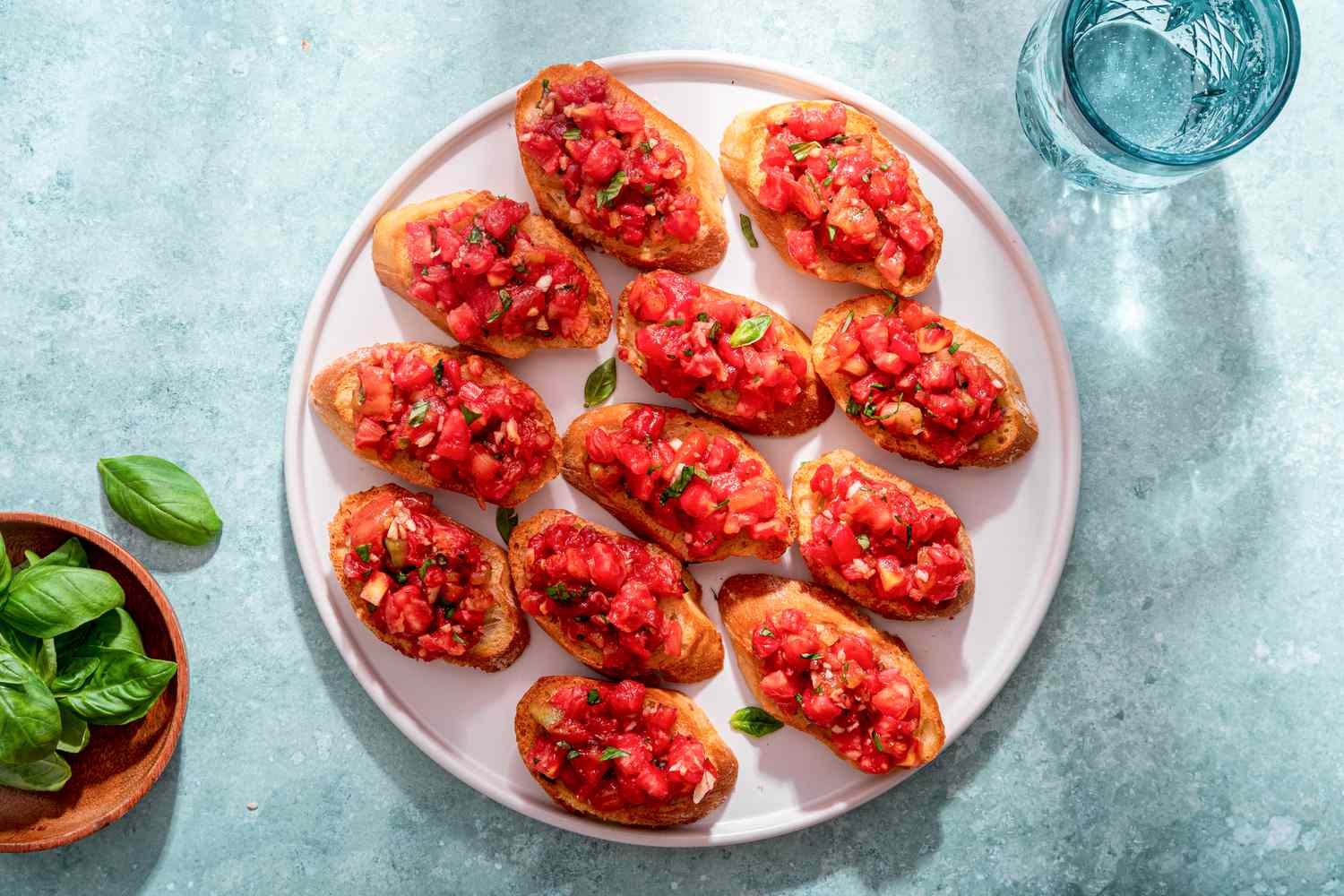 Plateful of bruschetta with tomato and basil next to a bowl of basil and a glass of water