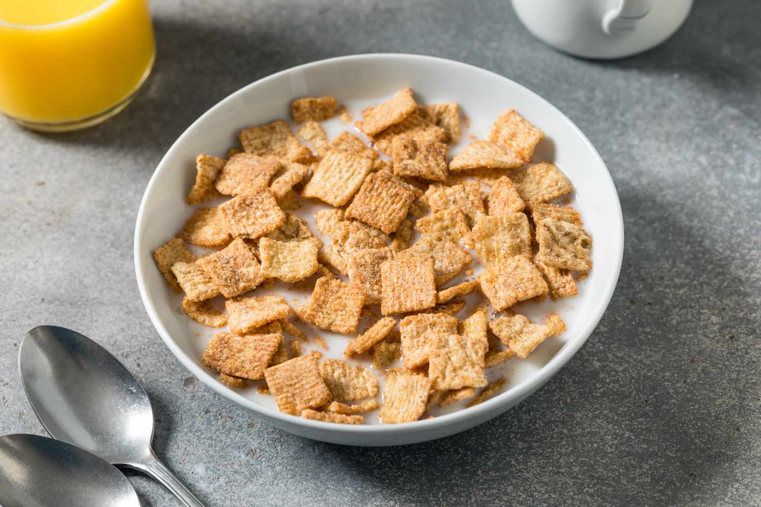 A bowl of cereal with milk on a table accompanied by spoons and a glass of orange juice