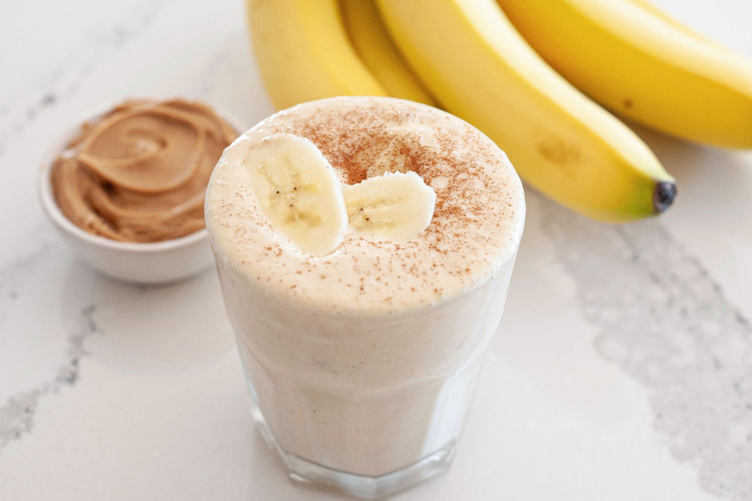 Simple peanut butter banana smoothie topped with sliced bananas and a bunch of bananas and a small container of peanut butter behind it.