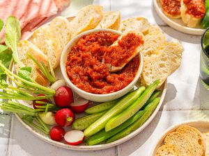 Side view of 2-ingredient honey chorizo spread in a bowl surrounded by radishes, cucumber spears and sliced baguette, with one baguette slice topped with the spread