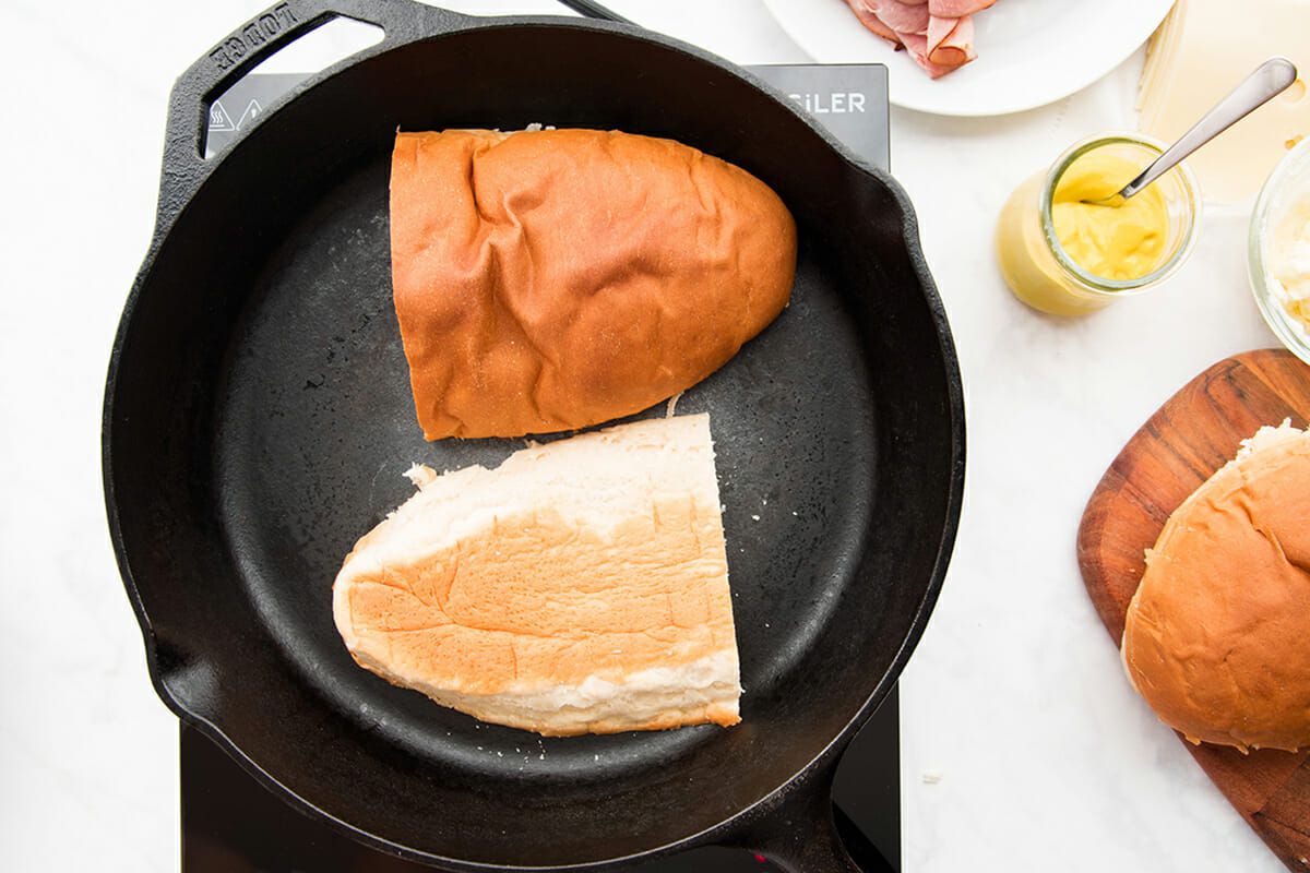 A cast iron skillet is set on a bunsen burner. Half of a loaf of french bread that has been lengthwise is being toasted to make the best cuban sandwich. A plate of sliced ham and sliced cheese is visible on the right side, along with a wooden cutting board and the other half of the french bread.