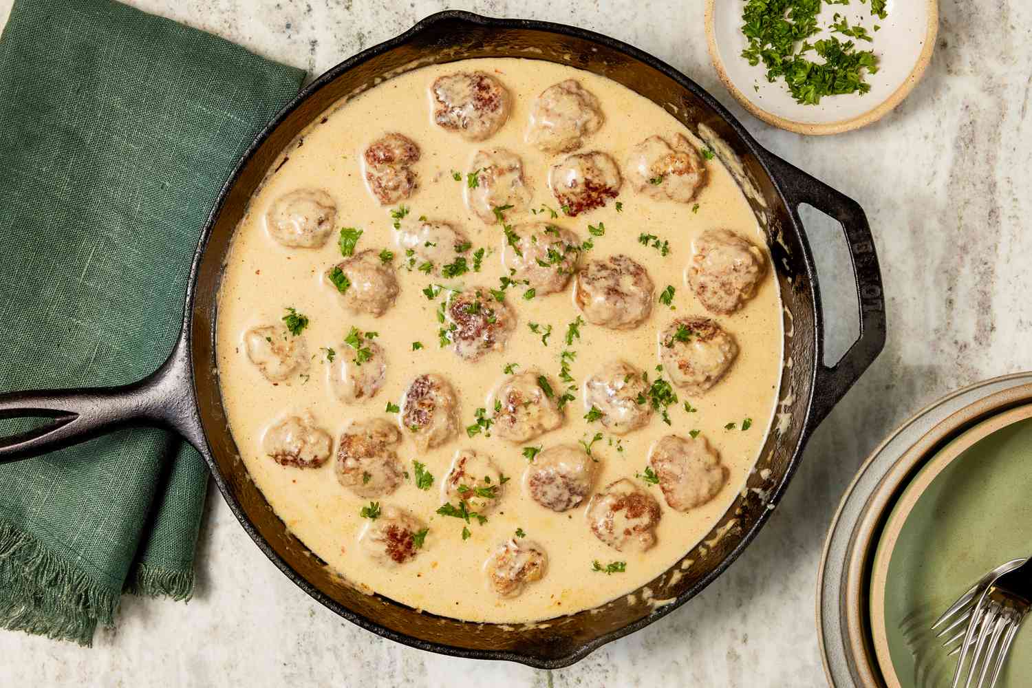 Cast iron skillet with copycat Ikea Swedish meatballs in gravy, and in the surroundings, a stack of plates with utensils, a small plate with minced herbs, and a sage table napkin 