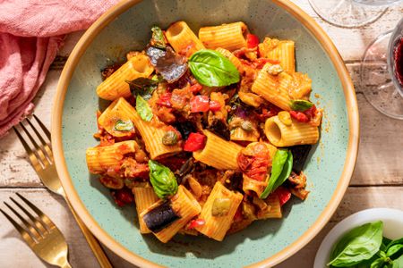 caponata pasta in a bowl (close up) next to a glass of wine, utensils on the table, and a small bowl of basil, all at a rustic table setting 