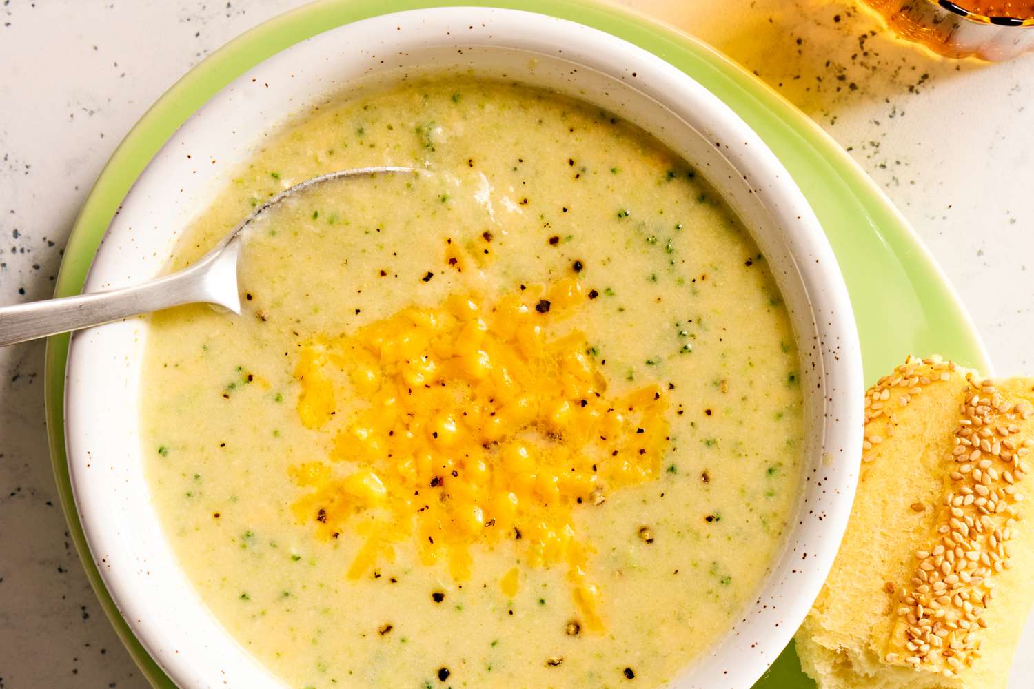 Overhead view of a bowl of broccoli cheddar soup with spoon on a plate with pieces of bread and next to a spoon and drink glass