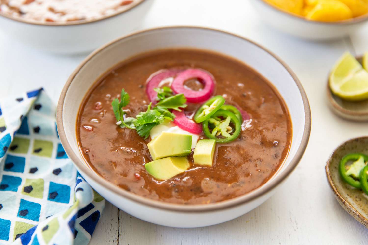A bowl of black bean soup, topped with avocado cubes, jalapeño slices, pickled onion and and cilantro