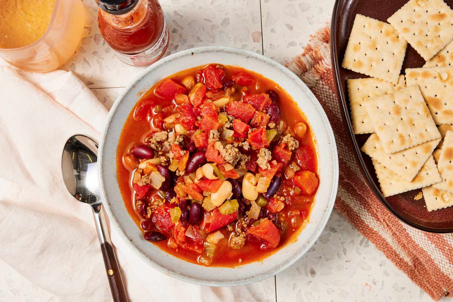 Bowl of copycat Wendy's chili at a table setting with a bowl of saltines on a brown and white kitchen towel, a bottle of tabasco, a drink, and a white table napkin