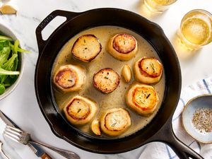 Fondant potatoes in a cast iron skillet at a table setting with a bowl of side salad, utensils on the counter, a bowl of pepper on a kitchen towel, and two glasses