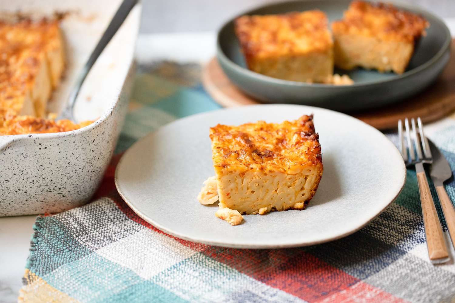 Slice of Trinidadian Macaroni Pie on a Plate on a Placemat, Surrounded by More Macaroni Pie in a Casserole Dish and a Shallow Bowl 