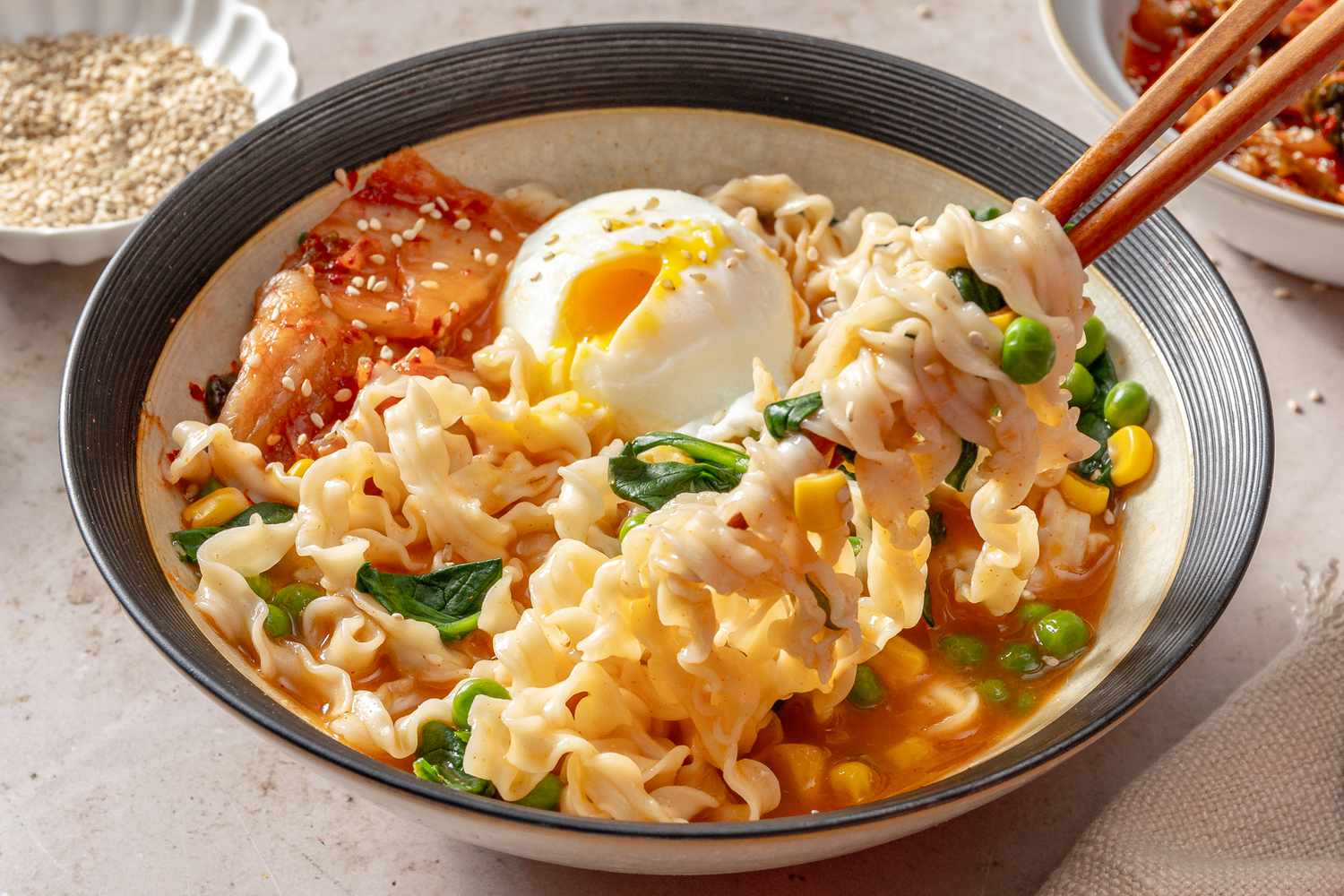A bowl of noodle soup with an egg, kimchi, vegetables, and chopsticks lifting noodles, garnished with sesame seeds