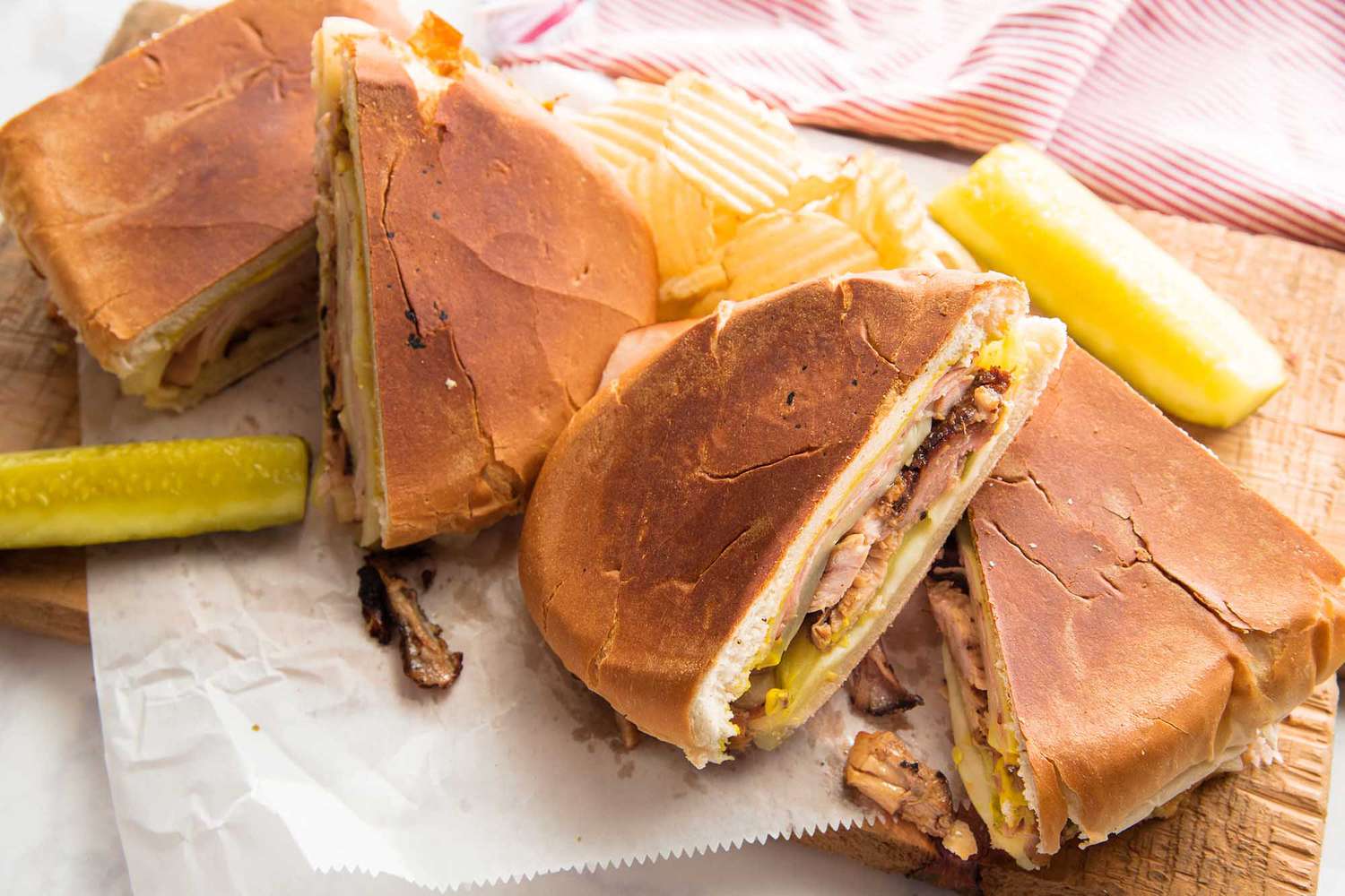 A grilled cuban sandwich is cut into quarters and resting on top of parchment paper and cutting board. Shredded pork and pickles are visible inside the sandwich. Pickles are on either side of the sandwich and a pink striped dish cloth is in the left hand corner.